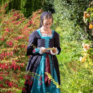 An image of Juliet Braidwood. They are standing among lush summer bushes, reading a paper in their hand. Their brown hair is visible under a black cap lined with white linen. They are wearing a black 16th century spiral laced bodice with a red sash and rich blue gown underneath. A white underdress with detailed blackwork embroidery can be seen at the neckline.