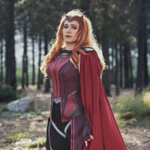 A picture of Lady Toxie. They ha ve red hair and are wearing a red headpiece. Their costume is two different shades of red with a bright red cape flowing behind them. They are looking directly into the camera with the sun beaming down from above. 