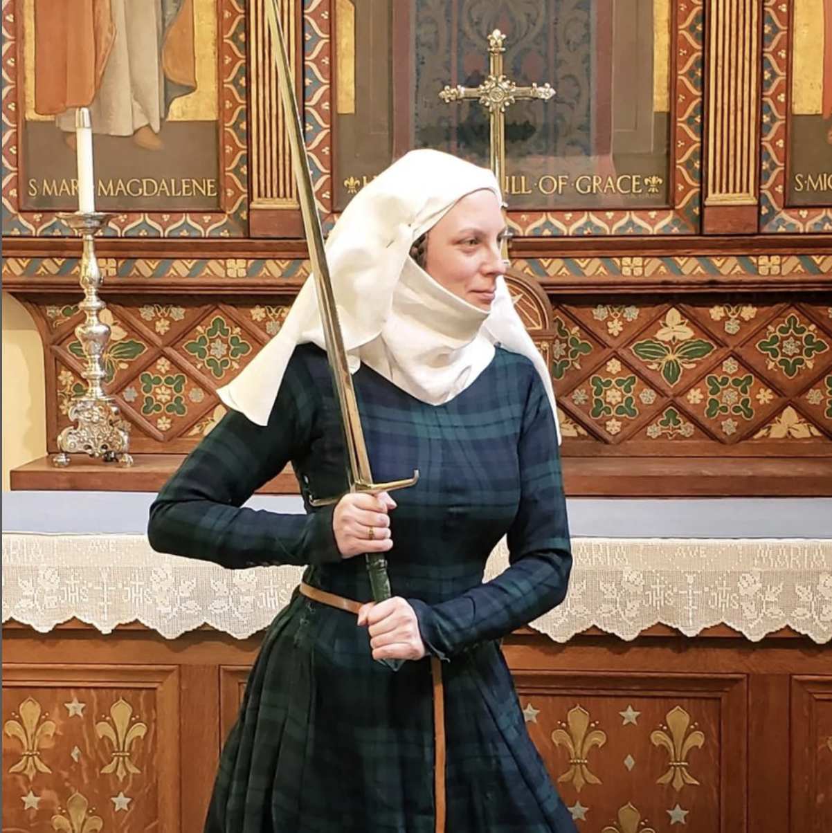 An image of Elisabeth Beattie. A human wearing a medieval white linen veil and vimple, green and blue medieval tartan dress, long belt, and holding a large sword with both hands. They are seemingly facing off with someone just out of frame. 