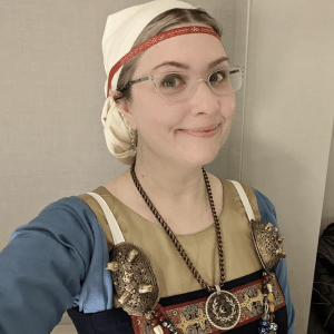 An image of Kerry Beckett, a person with glasses and blonde hair. They are wearing a headcovering, red headband with gold details, a beige and blue underdress and an apron dress with big tortoiseshell brooches, a large necklace and decorative details at the front. They are smiling at the camera. 