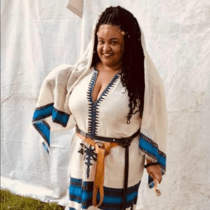 A picture of Jessica, smiling brightly at the camera. She is wearing a white linen shirt with bright blue and black trimmings at sleeve and hem. Black embroidery covers the front as well. Her long dark curls are worn loose and she wears a gold headpiece over her hair. 